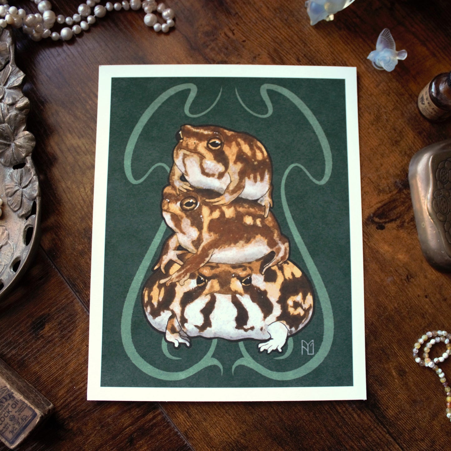 Illustration of a stack of three rain frogs on a green background with a wooden surface and jewelry around.