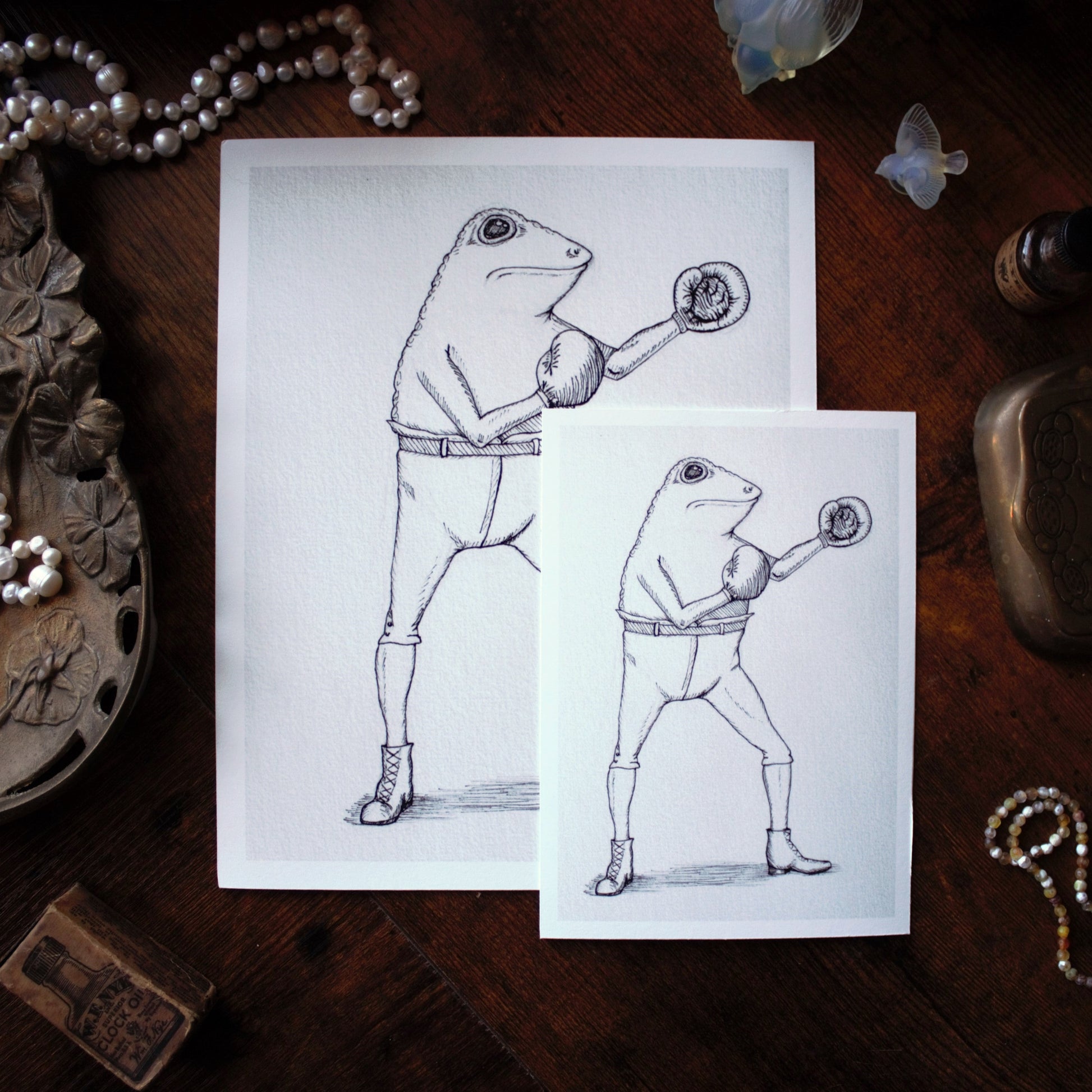 Two drawings of a frog in boxing gloves on a wooden surface with decorative items.