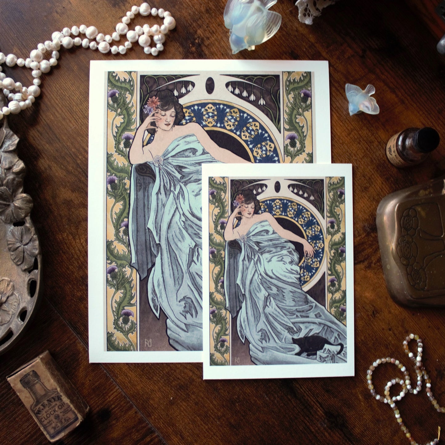 Two Art Nouveau-style prints featuring women in flowing dresses on a wooden surface with decorative elements.