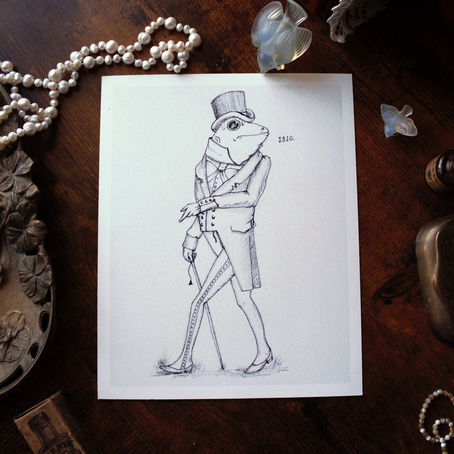 Drawing of a frog in formal attire on a wooden surface with decorative items.