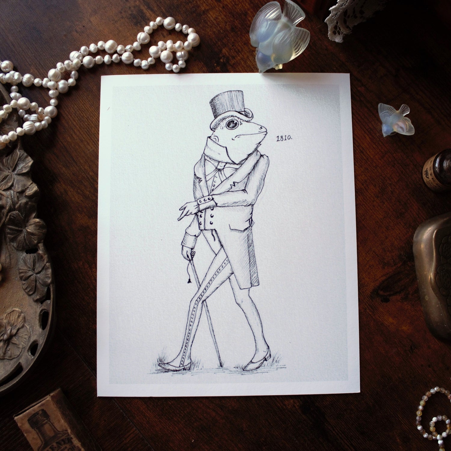 Drawing of a frog in formal attire on a wooden surface with decorative items.
