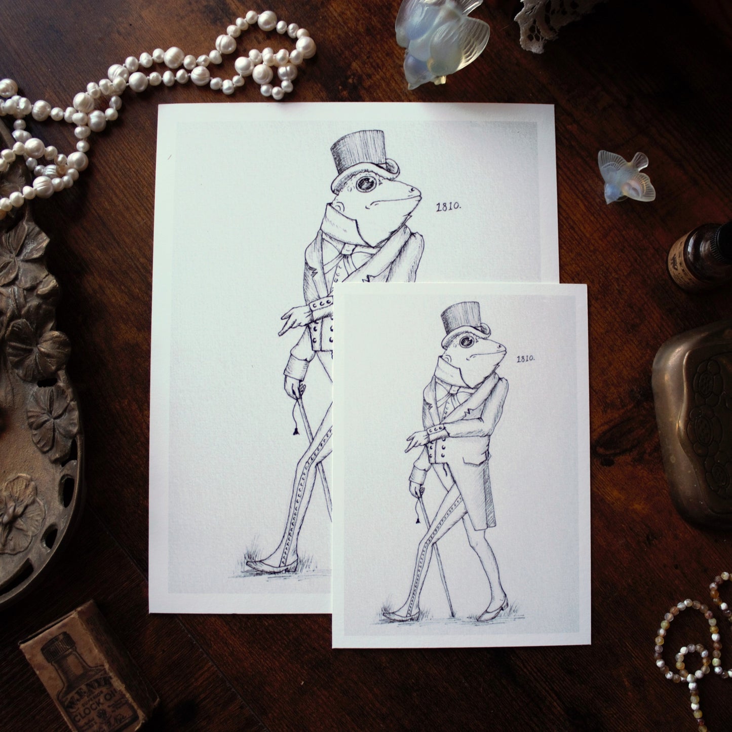 Two illustrations of a frog in formal attire on white paper, surrounded by decorative items on a wooden surface.