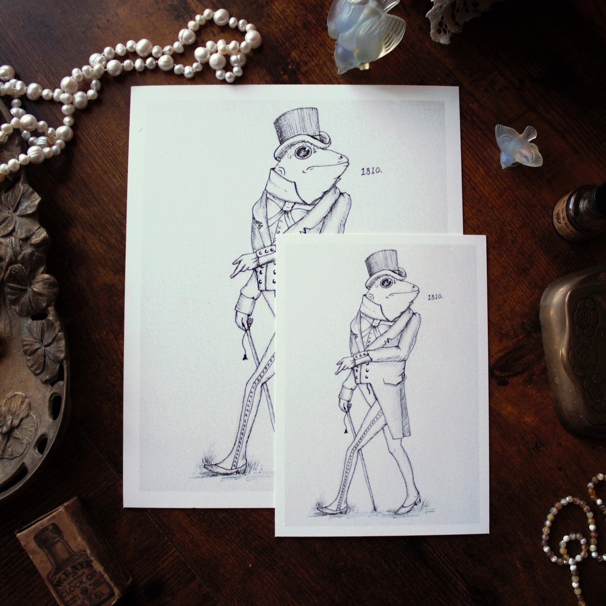 Two illustrations of a frog in formal attire on white paper, surrounded by decorative items on a wooden surface.