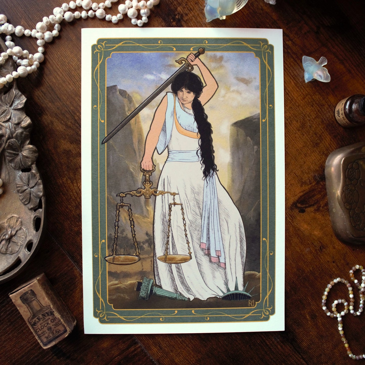 A print featuring a woman holding a sword and scales on a wooden surface with jewelry and a bottle.