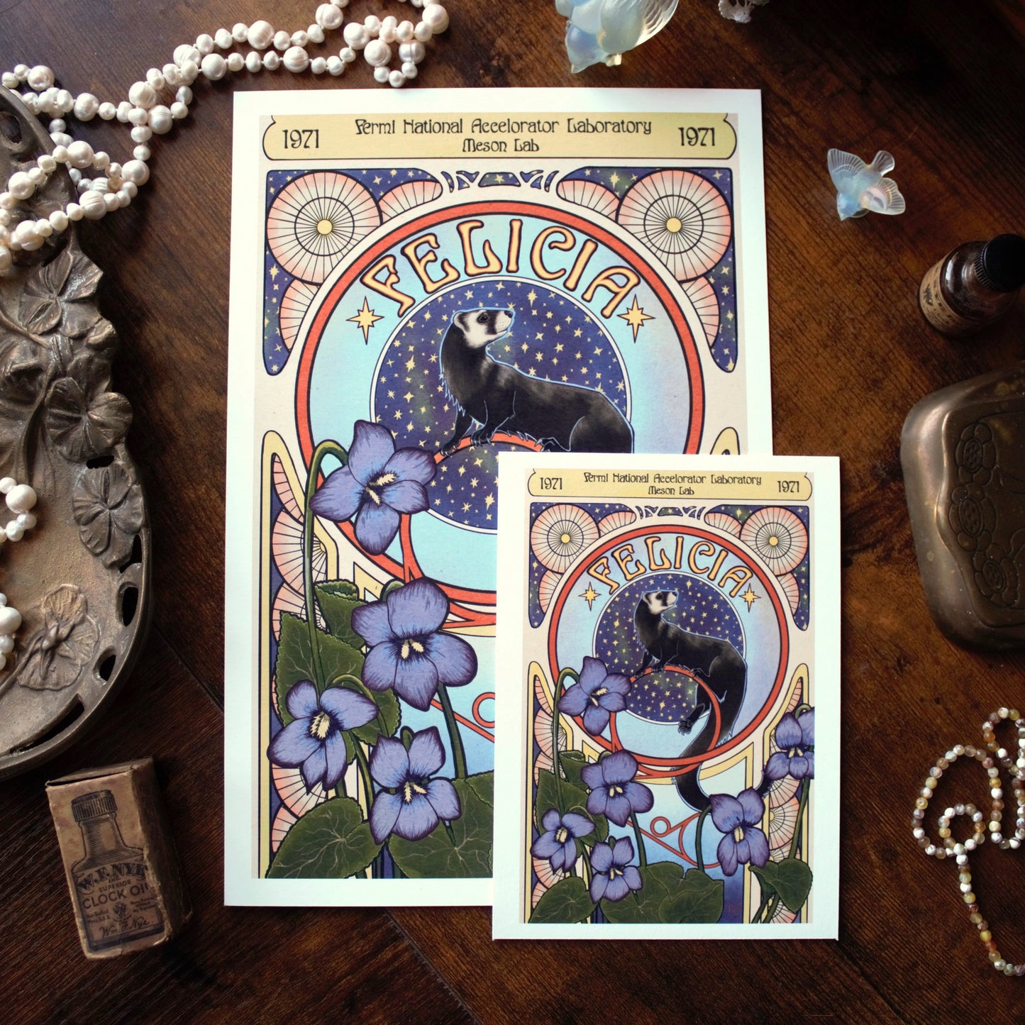 Two prints of an illustration of Felicia the ferret and flowers on a wooden surface with decorative items.